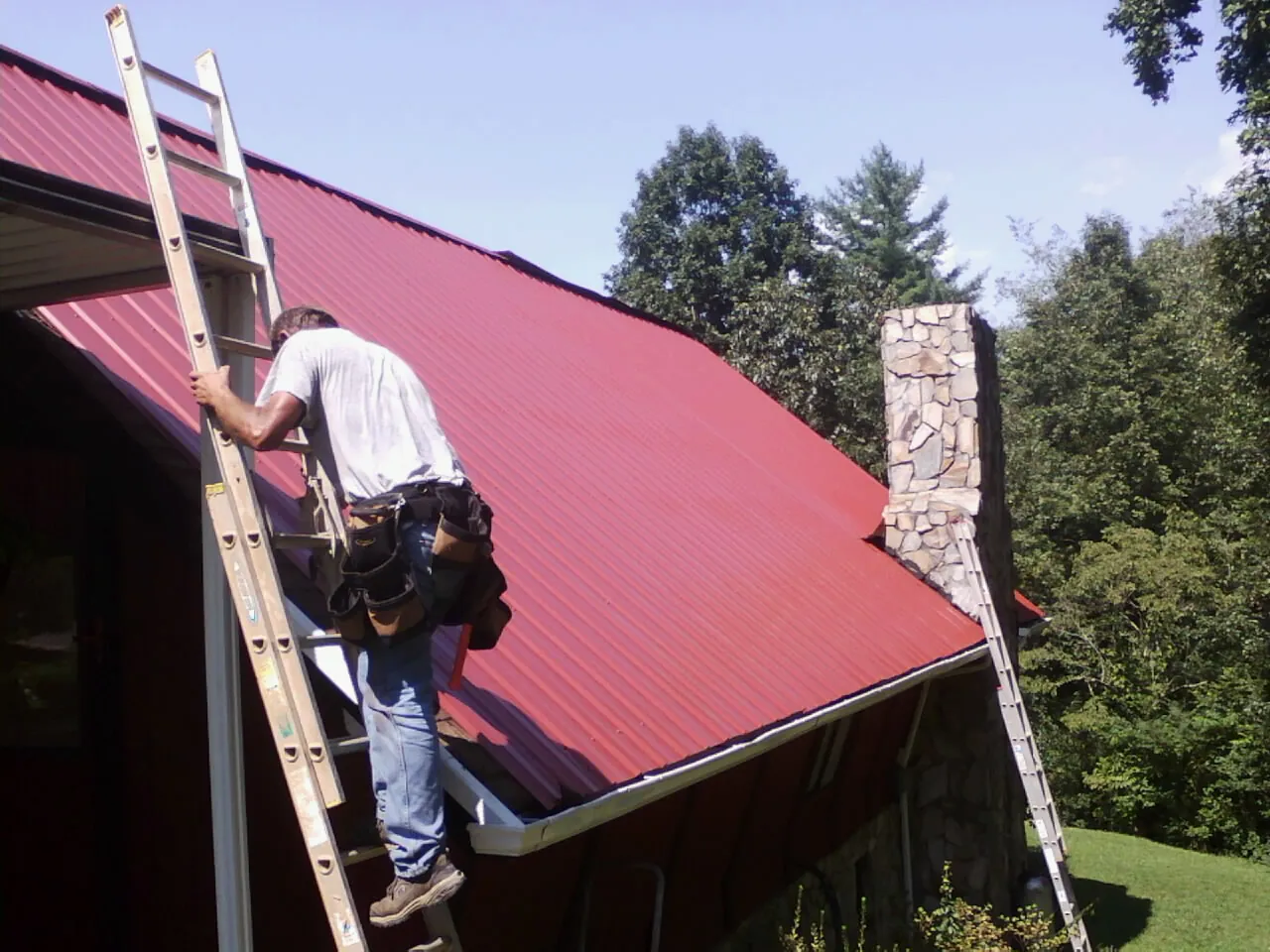 Roofer on a ladder beside a red metal roof and stone chimney — Soffit & Fascia Repair by Roofing Brothers in Wendover